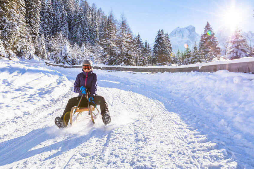 Cheerful girl riding a sled downhill on a snow covered sledge trail in a white sunny winter mountain landscape