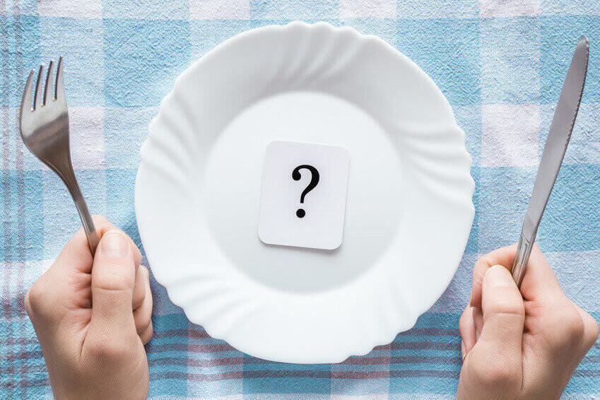 Woman's hands holding a fork and knife. Question mark in the clean, empty, white plate on the tablecloth. Starving people. Meal waiting concept. Top view.
