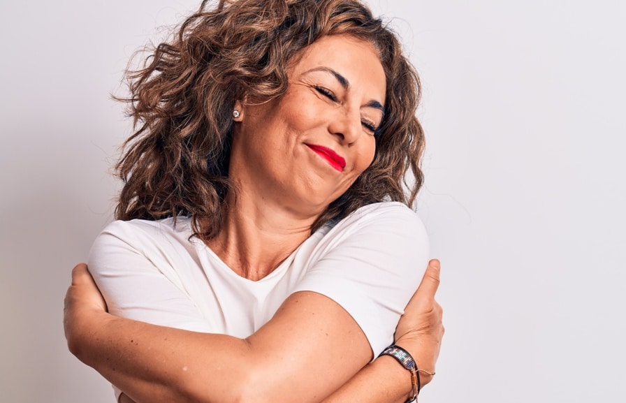 Middle age beautiful brunette woman wearing casual t-shirt standing over white background hugging oneself happy and positive, smiling confident. Self love and self care
