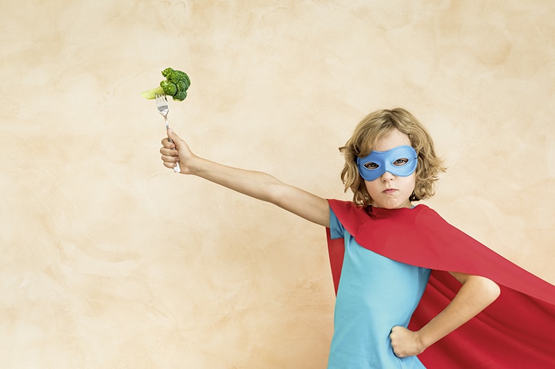 Superhero child eating superfood. Happy kid holding broccoli. Healthy eating and lifestyle concept. Green vegetarian food
