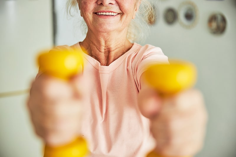 Close up of joyful old lady holding yellow dumbbells and smiling while having morning workout